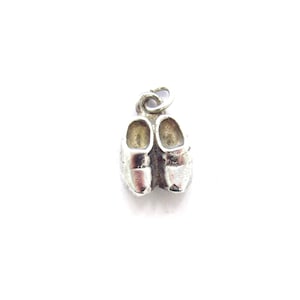May include: A small, silver-colored charm in the shape of a pair of clogs. The charm has a loop at the top for attaching to a necklace or bracelet. The clogs are detailed with visible grooves and a rounded toe.