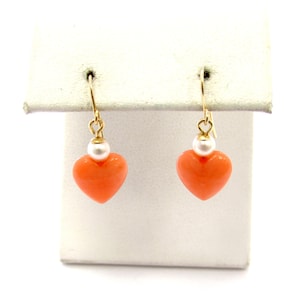 May include: A pair of heart-shaped earrings. Each earring features a vibrant orange heart pendant suspended from a gold-colored hook. A small white pearl sits above each heart. The earrings are displayed on a white surface.