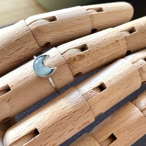 Könnte beinhalten: Ein silberner Halbmondring mit einem hellblauen Edelstein. Der Ring befindet sich auf einem hölzernen Handmodell.