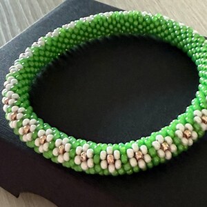 May include: A green and white beaded bracelet with a floral pattern. The bracelet is on a black background.