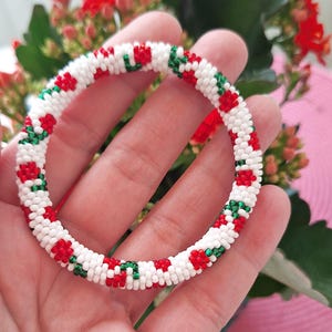 May include: A beaded bracelet with a repeating pattern of red and green floral designs on a white background. The bracelet is a circular shape and appears to be handmade. The bracelet is a piece of jewelry.