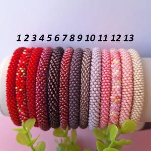 Könnte beinhalten: Dreizehn Perlenarmbänder in verschiedenen Farben, darunter Rot, Burgunder, Schwarz, Lila und Rosa. Die Armbänder werden auf einem weißen zylindrischen Ständer präsentiert, mit grünem Laub im Vordergrund. Die Zahlen 1 bis 13 sind darüber gedruckt.