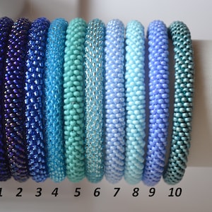 Puede incluir: Diez pulseras de cuentas en tonos de azul, turquesa y verde azulado, dispuestas en un degradado de azul oscuro a verde azulado claro. Las pulseras están numeradas del 1 al 10.