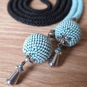 May include: A handmade necklace featuring a beaded design in black and light blue. The necklace has two light blue beaded spheres with silver-toned teardrop pendants. The necklace is displayed on a wooden surface.