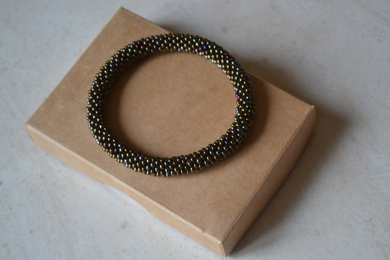 May include: A brown beaded bracelet with iridescent accents. The bracelet is on a brown cardboard box.