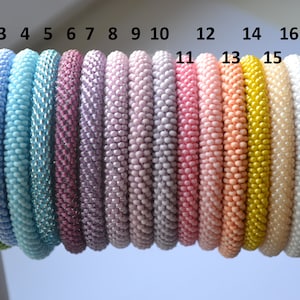 Può includere: Una fila di 17 braccialetti colorati con perline, numerati da 1 a 17. I braccialetti sono fatti di piccole perline rotonde di vari colori, tra cui verde, blu, viola, rosa, giallo e bianco.
