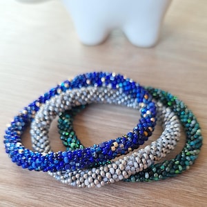 Könnte beinhalten: Drei Perlenarmbänder in Blautönen, Silber und Grün. Die Armbänder bestehen aus kleinen, runden Perlen und sind übereinander gestapelt.