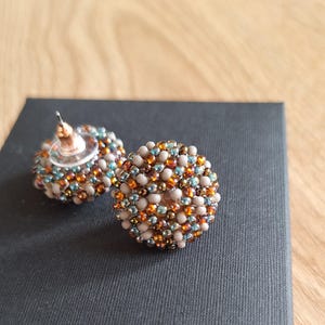 Könnte beinhalten: Ein Paar runde Perlenohrringe in einem braunen, beige und blauen Farbschema. Die Ohrringe bestehen aus kleinen Perlen, die zu einer Kugelform zusammengefügt sind.