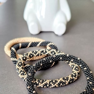 Könnte beinhalten: Vier Perlenarmbänder, zwei schwarz und zwei schwarz mit goldenen Akzenten. Die Armbänder sind übereinander gestapelt.