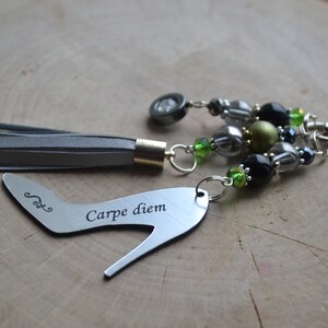 Puede incluir: Un charm de tacón alto plateado con la inscripción "Carpe diem" unido a un llavero plateado con un borla gris y una variedad de cuentas negras, verdes y plateadas.