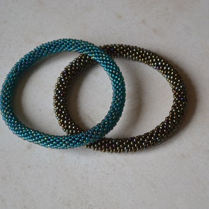 May include: Two beaded bracelets, one teal and one brown, with iridescent shimmer. The bracelets are made of small, round beads and are stacked on top of each other.