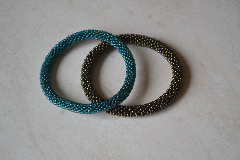 May include: Two beaded bracelets, one teal and one brown, with iridescent shimmer. The bracelets are made of small, round beads and are stacked on top of each other.