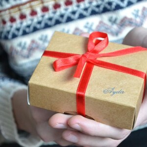 May include: A brown gift box with a red ribbon and bow. The box is tied with a red ribbon and has the word "Ayda" written on it in blue ink.