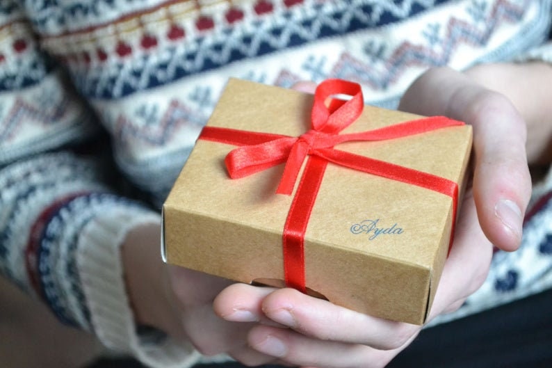 May include: A brown gift box with a red ribbon and bow. The box is tied with a red ribbon and has the word "Ayda" written on it in blue ink.