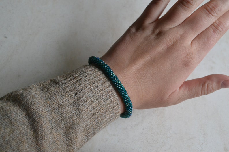 May include: A teal blue beaded bracelet with a spiral design. The bracelet is worn on a person's wrist.
