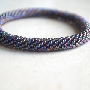 May include: A close-up of a multi-coloured beaded bracelet. The bracelet is made of small, iridescent beads in shades of purple, blue, and green. The beads are arranged in a spiral pattern, creating a textured and eye-catching design.