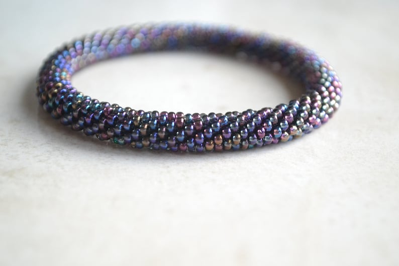 May include: A close-up of a multi-coloured beaded bracelet. The bracelet is made of small, iridescent beads in shades of purple, blue, and green. The beads are arranged in a spiral pattern, creating a textured and eye-catching design.