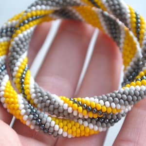 May include: A close-up of a beaded bracelet with a yellow, white, and gray color scheme. The bracelet is made of small, round beads that are woven together in a spiral pattern.