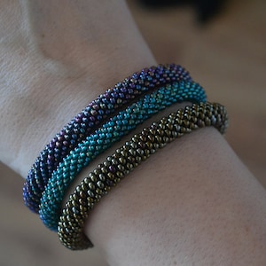 May include: Three beaded bracelets stacked on a wrist. The bracelets are made of small, iridescent beads in shades of purple, teal, and brown.