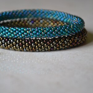 May include: Three beaded bracelets stacked on top of each other. The top bracelet is teal blue, the middle bracelet is gold, and the bottom bracelet is brown.