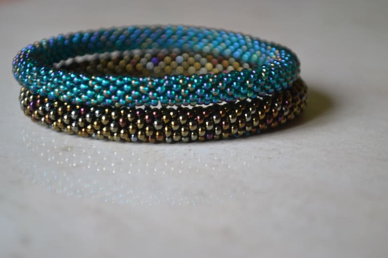 May include: Three beaded bracelets stacked on top of each other. The top bracelet is teal blue, the middle bracelet is gold, and the bottom bracelet is brown.