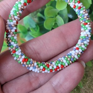 May include: A beaded bracelet with a spiral design. The bracelet features small, colorful beads in green, white, red, and blue, interspersed with gold-colored beads. The bracelet is held in a hand, showcasing its circular form and intricate pattern.