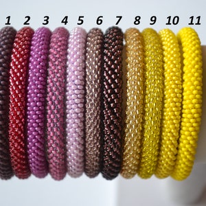 Könnte beinhalten: Elf Perlenarmbänder in verschiedenen Farben, darunter Lila, Rosa, Braun, Gold und Gelb. Die Armbänder sind von 1 bis 11 nummeriert.