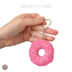 May include: A pink crocheted donut keyring with green and white sprinkles. The donut is attached to a silver heart-shaped keyring.
