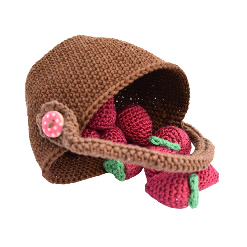 Puede incluir: Una cesta de crochet marr&oacute;n llena de fresas de crochet rojas. La cesta tiene un asa marr&oacute;n y un bot&oacute;n rosa con lunares blancos.