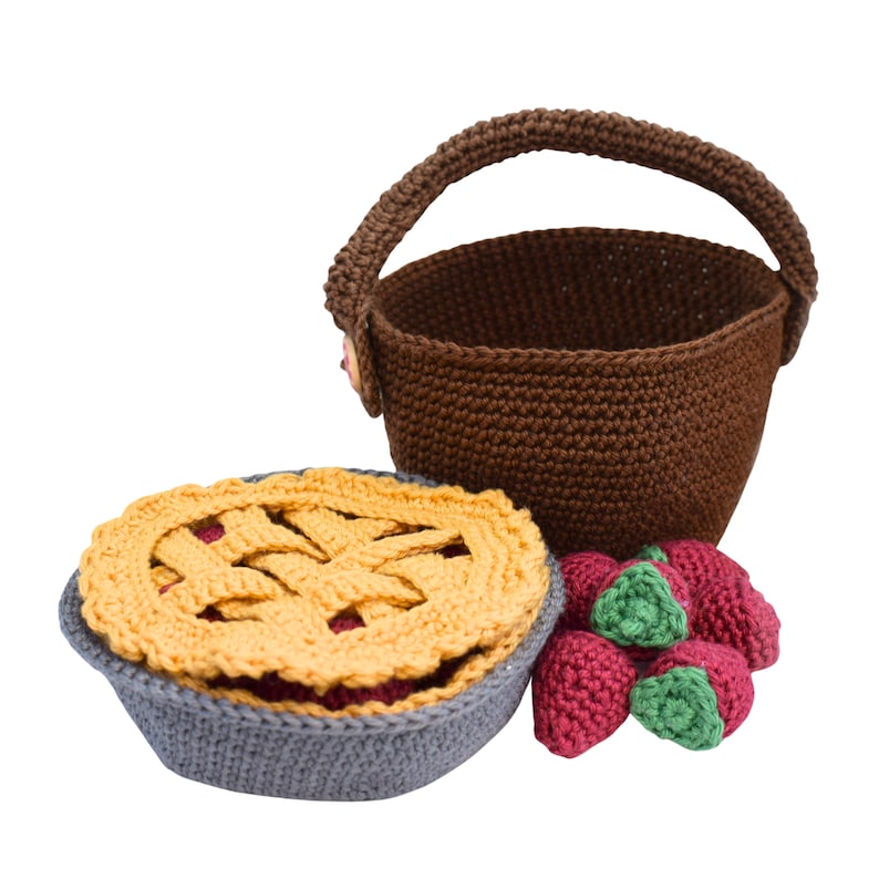 Puede incluir: Una cesta de crochet marr&oacute;n con asa, una tarta de crochet con costra amarilla y relleno rojo, y un grupo de fresas rojas de crochet con hojas verdes.