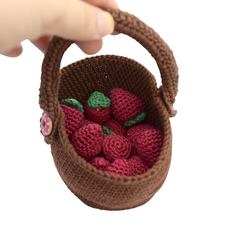 Puede incluir: Una cesta de crochet marr&oacute;n llena de fresas rojas de crochet. La cesta tiene un asa marr&oacute;n y un bot&oacute;n rosa.