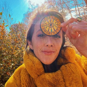May include: A woman wearing a mustard yellow faux fur coat holds a round sticker with the words "INCREASED AWARENESS" around an eye design. The background features fall foliage and a clear blue sky.