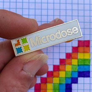 Peut inclure: Une épingle en argent avec un fond blanc et le mot "Microdose" en lettres noires. L'épingle a un motif coloré de quatre carrés dans le coin supérieur gauche.