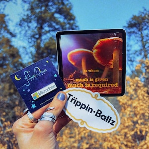 Pode incluir: Uma mão segurando um adesivo que diz "Trippin-Ballz" com um desenho de cogumelo. O adesivo está em cima de um cartão com o texto "a quem muito é dado, muito é pedido" e uma imagem de cogumelo. A mão também está segurando um cartão pequeno com o texto "Future Dawn Designs" e um alfinete que diz "Microdose".
