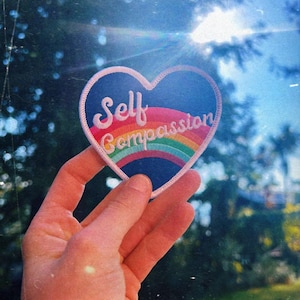 Pode incluir: Um remendo em forma de coração com um design de arco-íris e o texto "Self Compassion" em letras brancas.