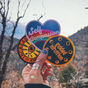 Pode incluir: Uma coleção de patches bordados com mensagens inspiradoras. Um patch tem formato de coração com "Self Compassion" sobre um arco-íris. Outro diz "Increase Awareness" com um design de olho. O terceiro diz "Guided Intuition".