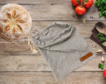 May include: A white and gray striped linen bread bag with a brown leather tag. The bag is sitting on a wooden surface with a loaf of bread and other kitchen items.