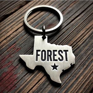 May include: Silver Texas-shaped keychain with a black inscription that reads "FOREST" and a silver star.