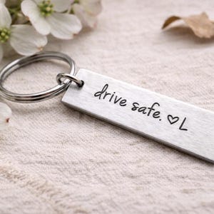 May include: A rectangular silver keychain with the words "drive safe. ♡ L" engraved in black. The keychain is attached to a silver ring. White flowers and a dried leaf are in the background.