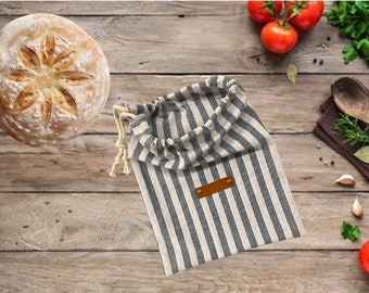 May include: A striped fabric bread bag with a brown leather label. The bag is on a wooden surface with a loaf of bread and tomatoes.