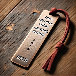 May include: A copper bookmark with a quote that reads "One chapter ends, another begins." The bookmark has a small tree design and the name "Beth" engraved on it. It is attached to a red leather cord.
