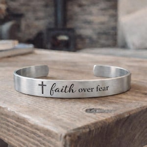 May include: A silver-colored cuff bracelet with the words "faith over fear" and a cross symbol engraved on the front. The bracelet is displayed on a rustic wooden table, showcasing its simple yet meaningful design.