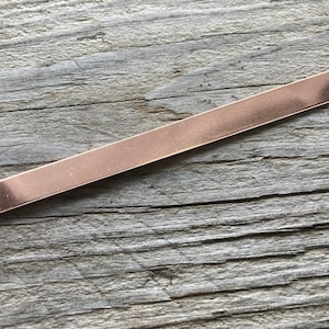 May include: A long, thin, flat piece of copper metal with rounded edges. The metal has a warm, reddish-brown color.