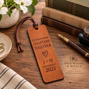 May include: A rectangular tan leather bookmark with a dark brown leather cord loop. The bookmark is engraved with the text TO MANY MORE CHAPTERS TOGETHER, a heart symbol, the initials J + M, and the date SEPTEMBER 23 2023 in a dark font.
