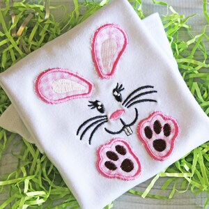 May include: White baby onesie with a pink and white bunny face design. The bunny face features pink ears, a pink nose, black whiskers, and two brown paw prints. The onesie is folded and placed on a bed of green Easter grass.