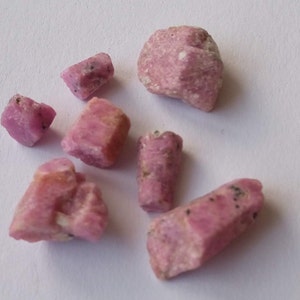 Puede incluir: Una colección de siete cristales de rubí rosa de diferentes tamaños y formas. Los cristales son ásperos y tienen un aspecto natural sin pulir.