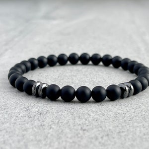 May include: A black beaded bracelet with matte finish beads. The bracelet has a few silver-coloured accent beads. The bracelet is displayed on a light grey surface.