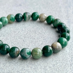 May include: A beaded bracelet featuring round, polished beads in shades of green and off-white. The beads are strung together to form a circular shape, showcasing variations in color and texture. The bracelet is a piece of jewelry.