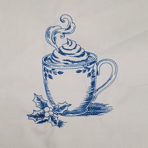 May include: Blue embroidered design of a coffee cup with whipped cream and steam. The cup has a handle and is decorated with floral patterns. A sprig of holly with berries is next to the cup. The design is on a white background.