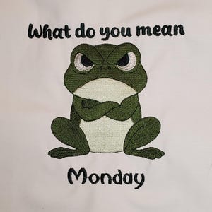 May include: White fabric featuring an embroidered design of a grumpy green frog with crossed arms. The text "What do you mean" is above the frog, and "Monday" is below.